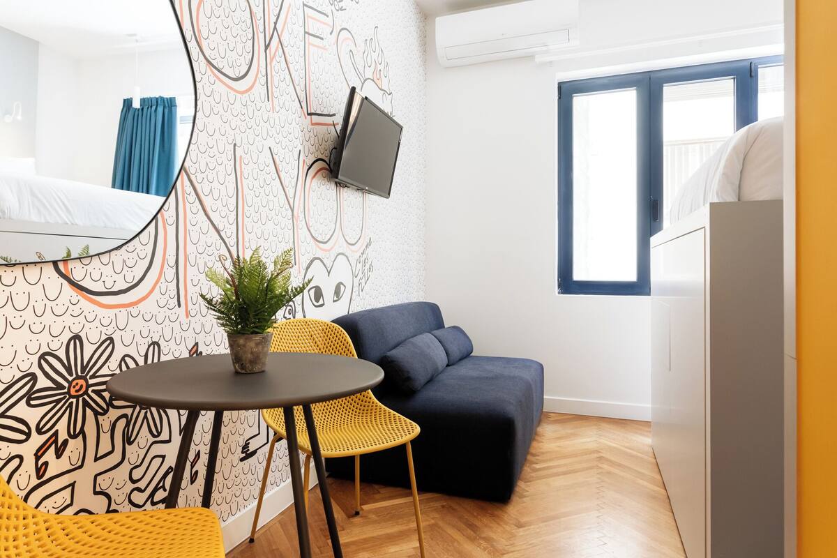 Un espacio habitable compacto con un sofá, una mesita redonda con una planta en maceta y un televisor montado en la pared.