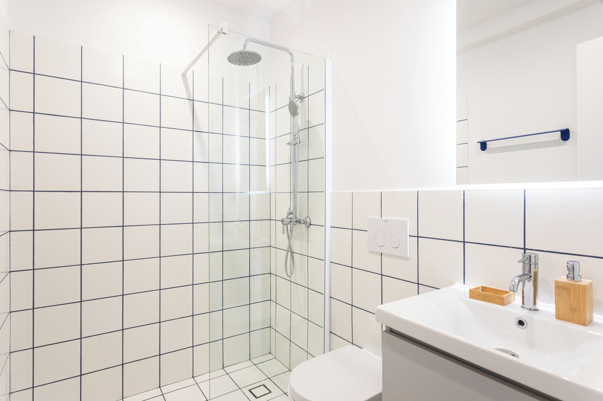 Et moderne badeværelse med en glasbrusekabine, hvide fliser og en hvid vask med en træ sæbeskål.