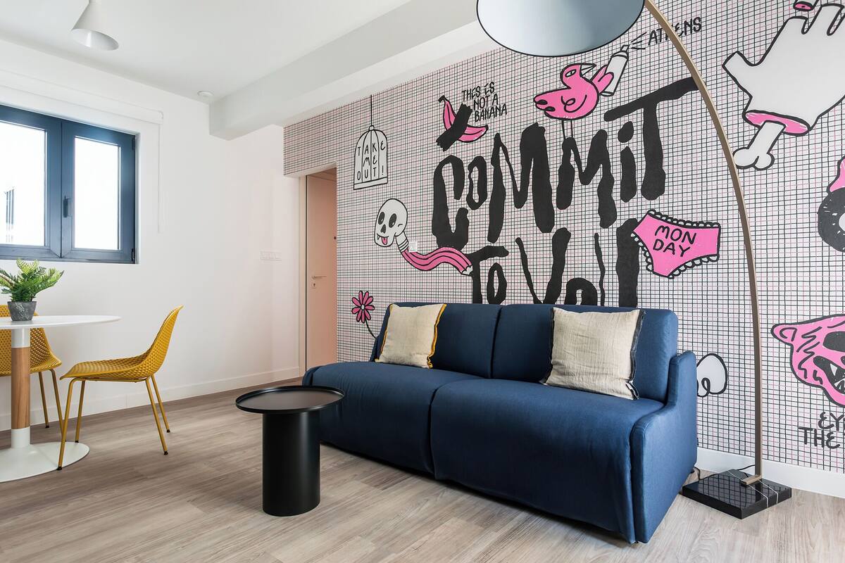 Una sala de estar moderna con un sofá azul, una mesa de centro negra redonda y una silla amarilla. Se aprecia un mural grande con diseños abstractos y texto.