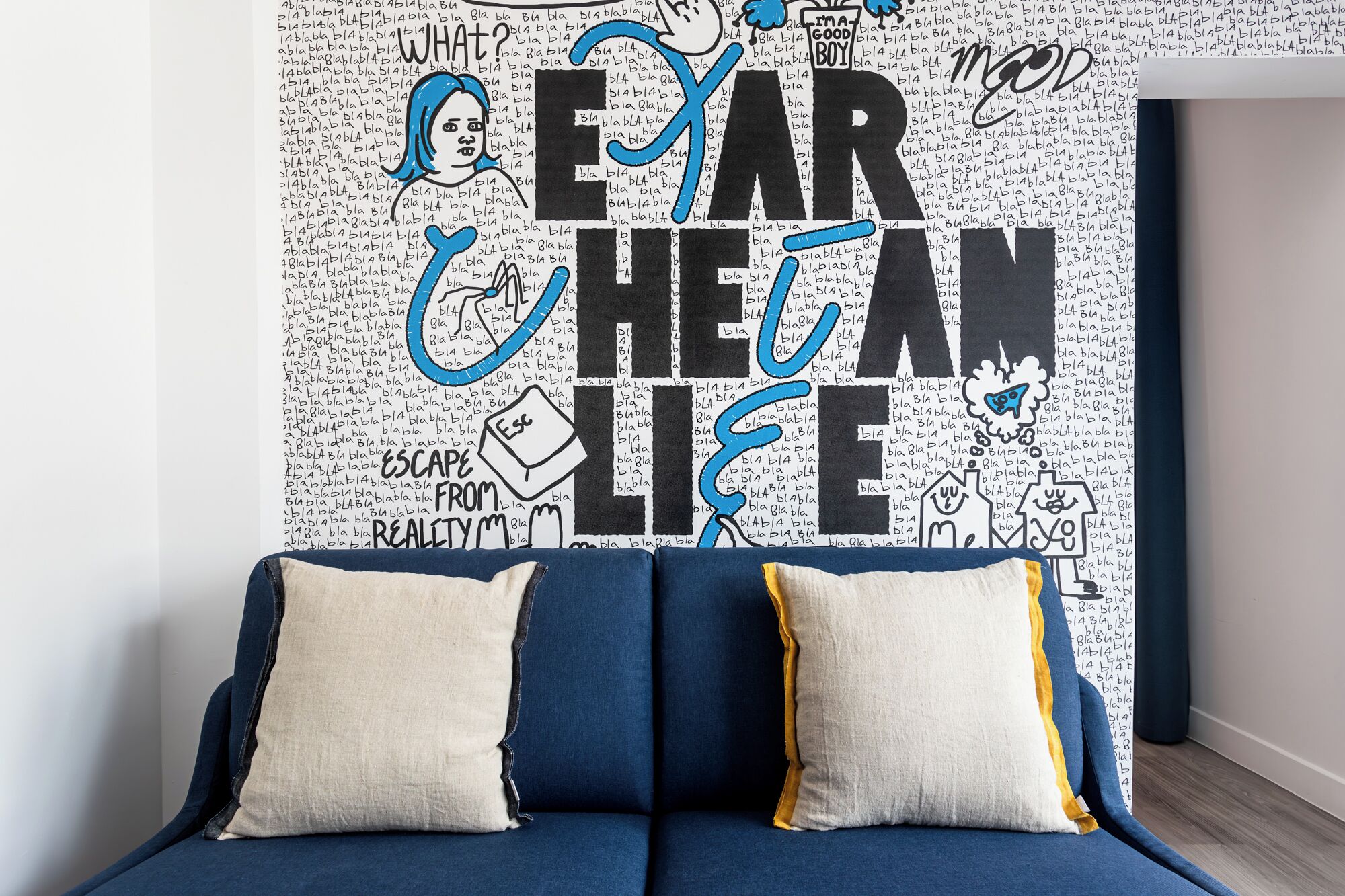 A room with a blue sofa and two pillows, a wall with a large, colorful mural, and a door to the right.