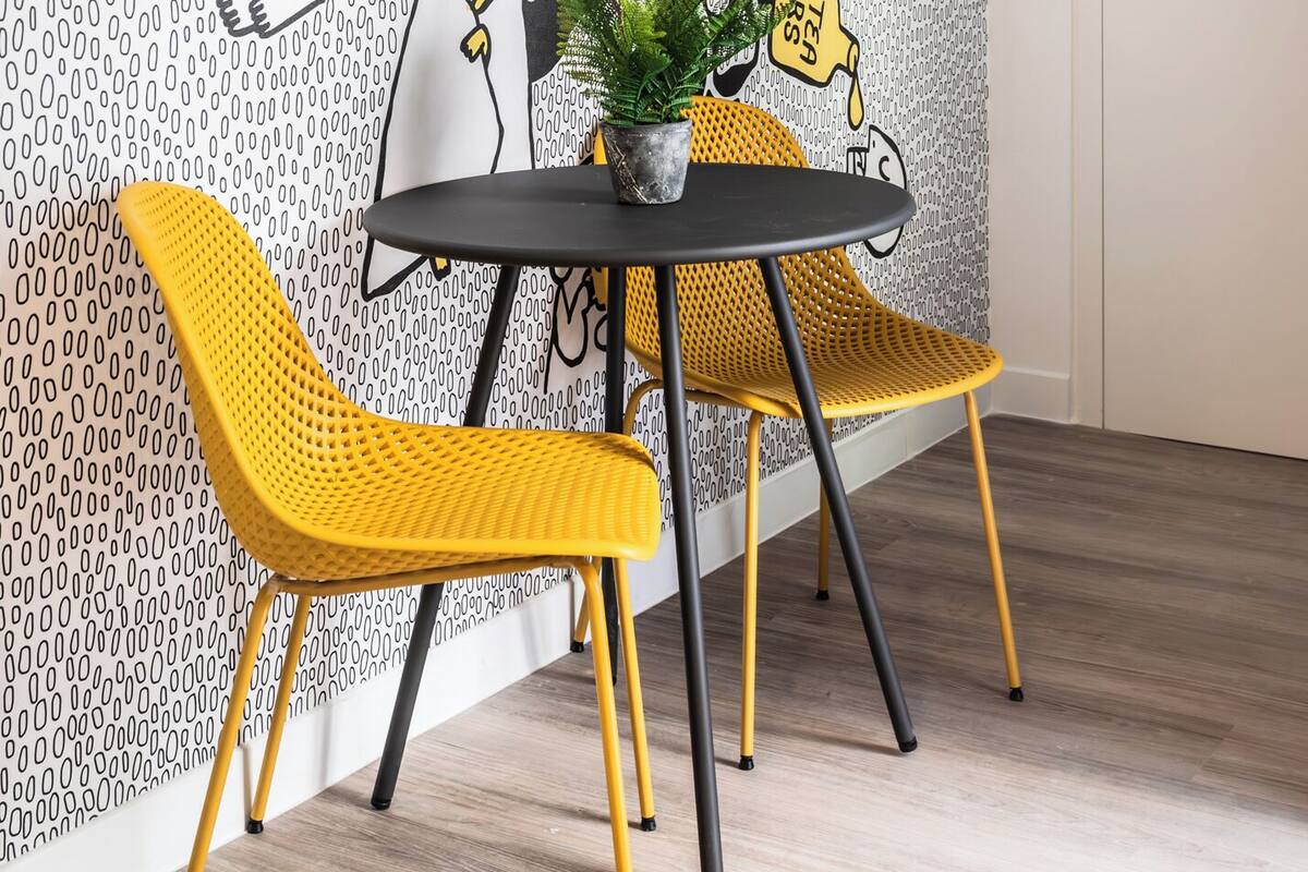 Una habitación con un papel tapiz estampado, una mesa redonda con dos sillas amarillas, una planta en maceta y un espejo grande.