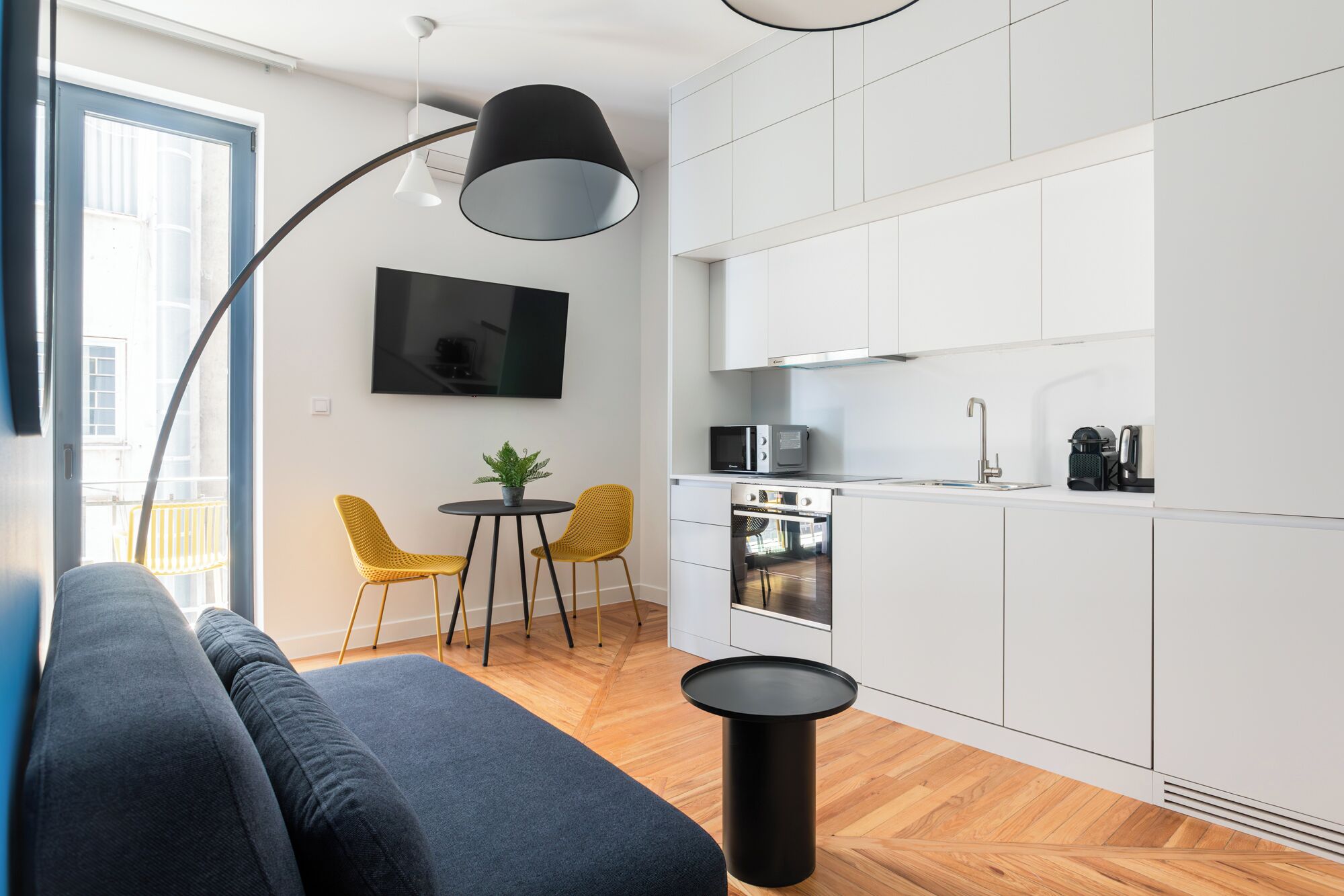A modern kitchen with white cabinets, a microwave, and a stove. There is a dining area with a round table and two yellow chairs. A black floor lamp and a black table are also visible.