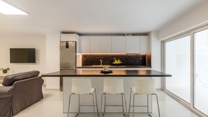 A modern kitchen with a black backsplash, white cabinets, and a large window. There are three white bar stools and a brown sofa. - Kookaki by Zoia Living (Athens)
