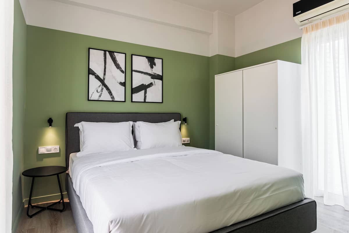 A neatly made bed in a room with green walls, two abstract paintings, a wardrobe, and a bedside table with a lamp.