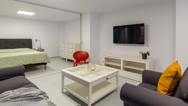 Un dormitorio moderno con cama, una mesita de centro de vidrio, una silla roja, un televisor y un aparador blanco.