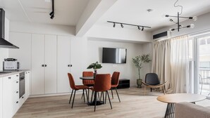 A modern kitchen with white cabinets, a built-in microwave, and a stove. There is a dining area with a round table and orange chairs. A TV is mounted on the wall, and a plant is on the table. - Kookaki by Zoia Living (Athens)
