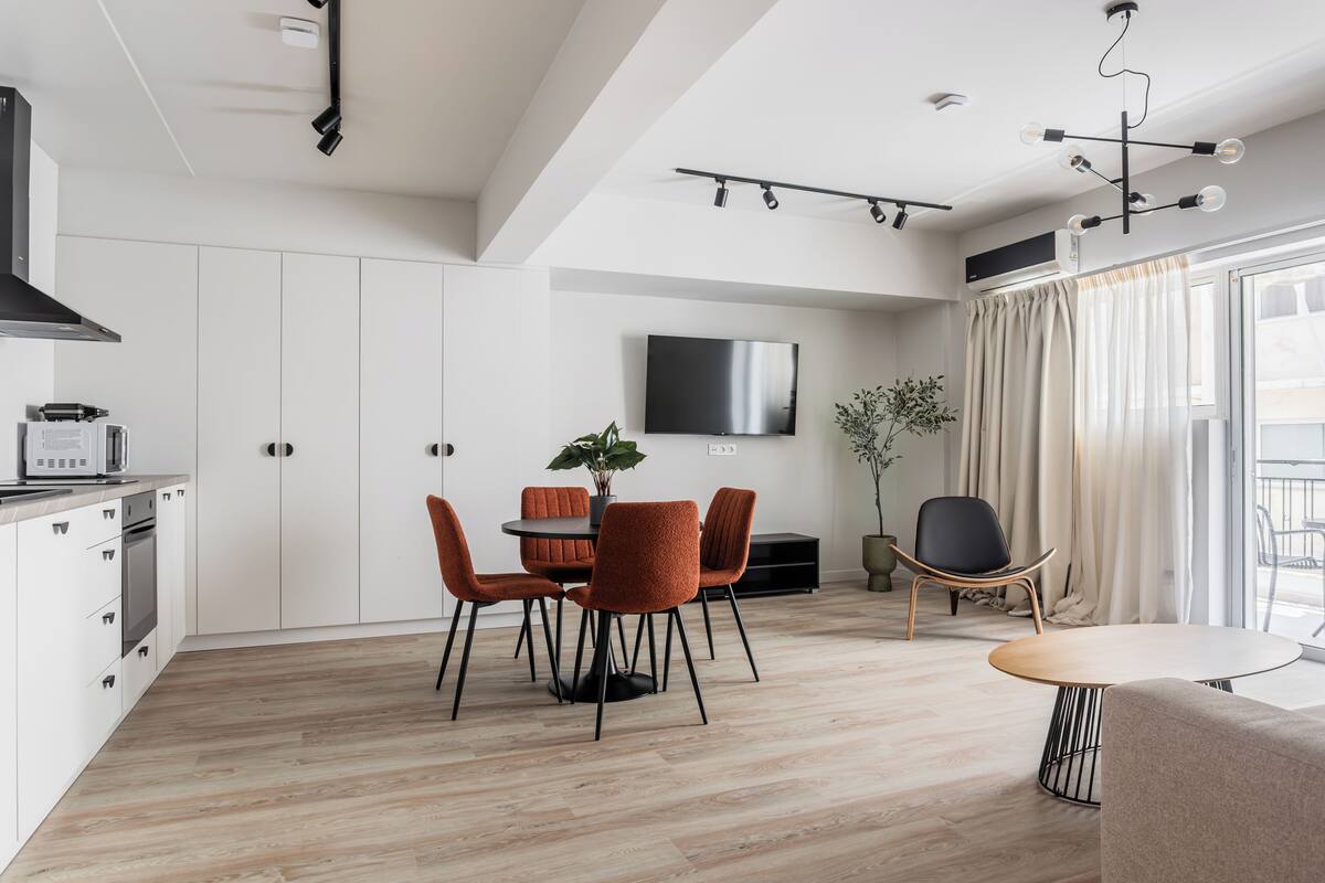 A modern kitchen with white cabinets, a built-in microwave, and a stove. There is a dining area with a round table and orange chairs. A TV is mounted on the wall, and a plant is on the table.