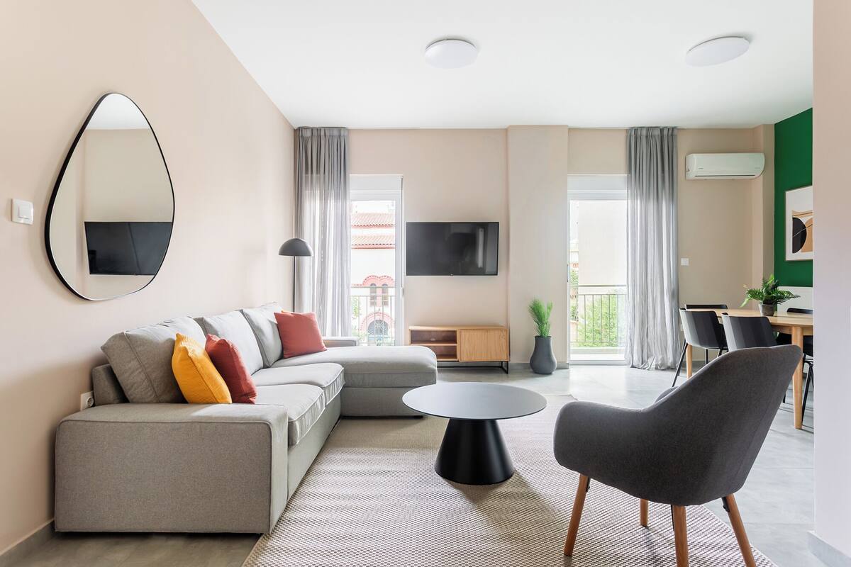 A modern living room with a grey sofa, a round coffee table, and a grey armchair. There is a TV mounted on the wall, a wooden cabinet, and a balcony with a view.