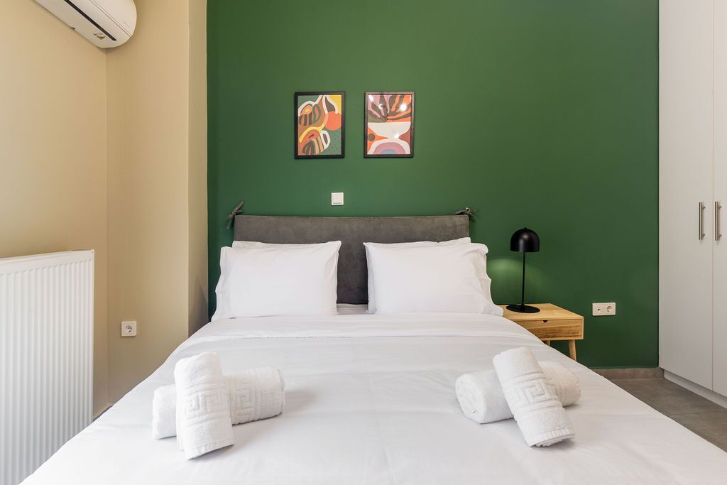 A neatly made bed with white linens and towels, a grey headboard, and two abstract paintings on a green wall.