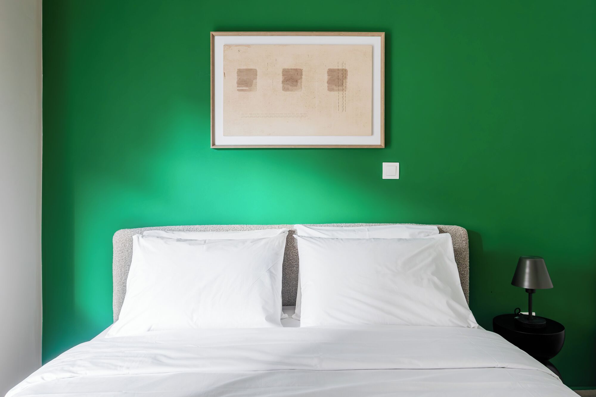 Een kamer met een groene wand, een lijst met foto, een bed met witte beddengoed en een nachtkastje met een lamp.
