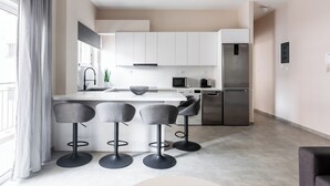 A modern kitchen with white cabinets, a stainless steel refrigerator, a microwave, and a sink. There are three grey bar stools around the kitchen island. - Snug by Zoia Living (Athens)