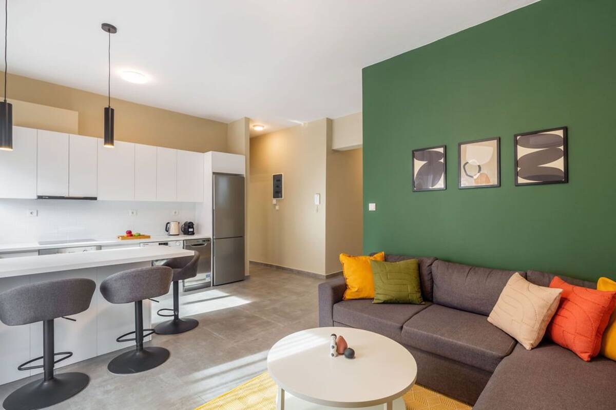 A modern apartment with a kitchen, dining area, and living room. It features a green accent wall, a gray sofa with colorful cushions, and a white coffee table.