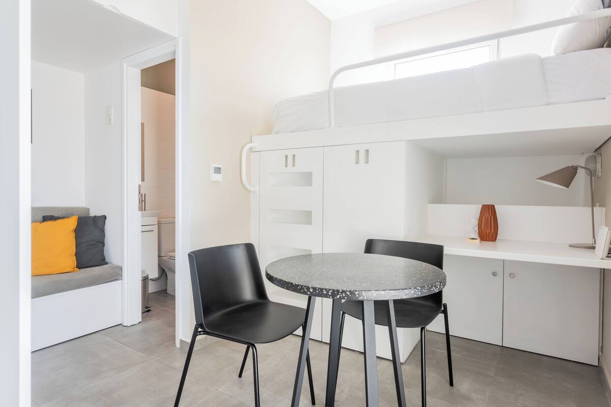 A compact room with a bunk bed, a small kitchenette, a round table with two chairs, and a sofa with cushions.