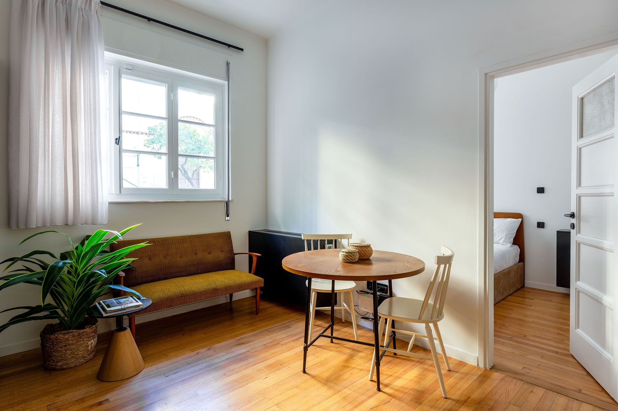 Una habitación acogedora con suelo de madera, una mesa redonda con sillas, un sofá, una planta en maceta y una ventana con cortinas.