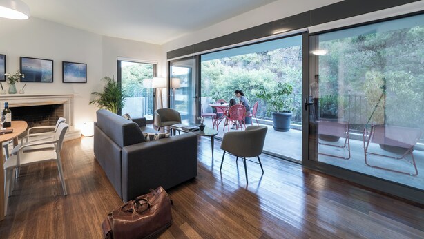 A modern living room with a fireplace, a sofa, a dining area, and large sliding glass doors leading to an outdoor patio. - The Hill by Zoia Living (Athens)