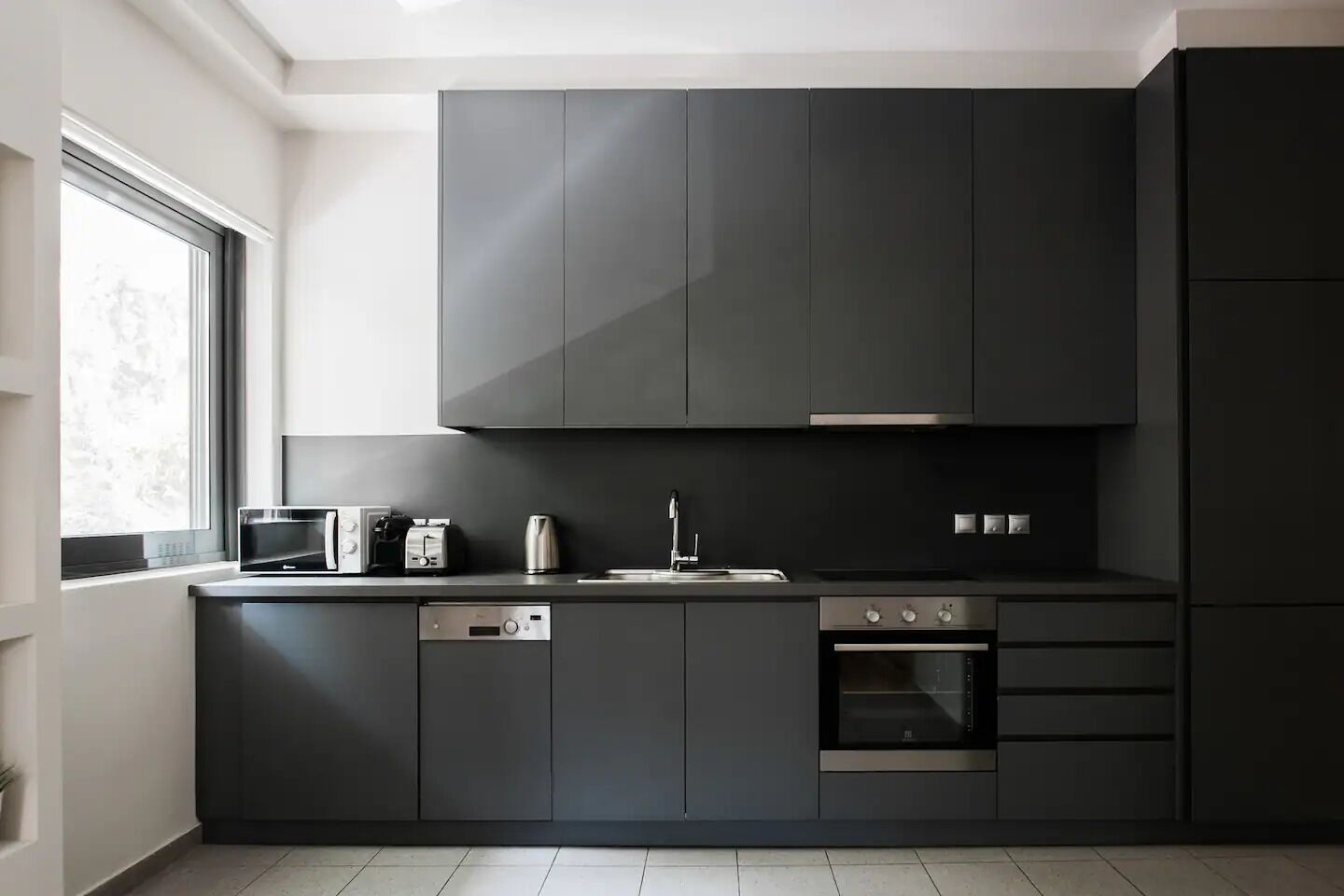 A modern kitchen with dark cabinetry, a built-in oven, a dishwasher, and a window providing natural light.