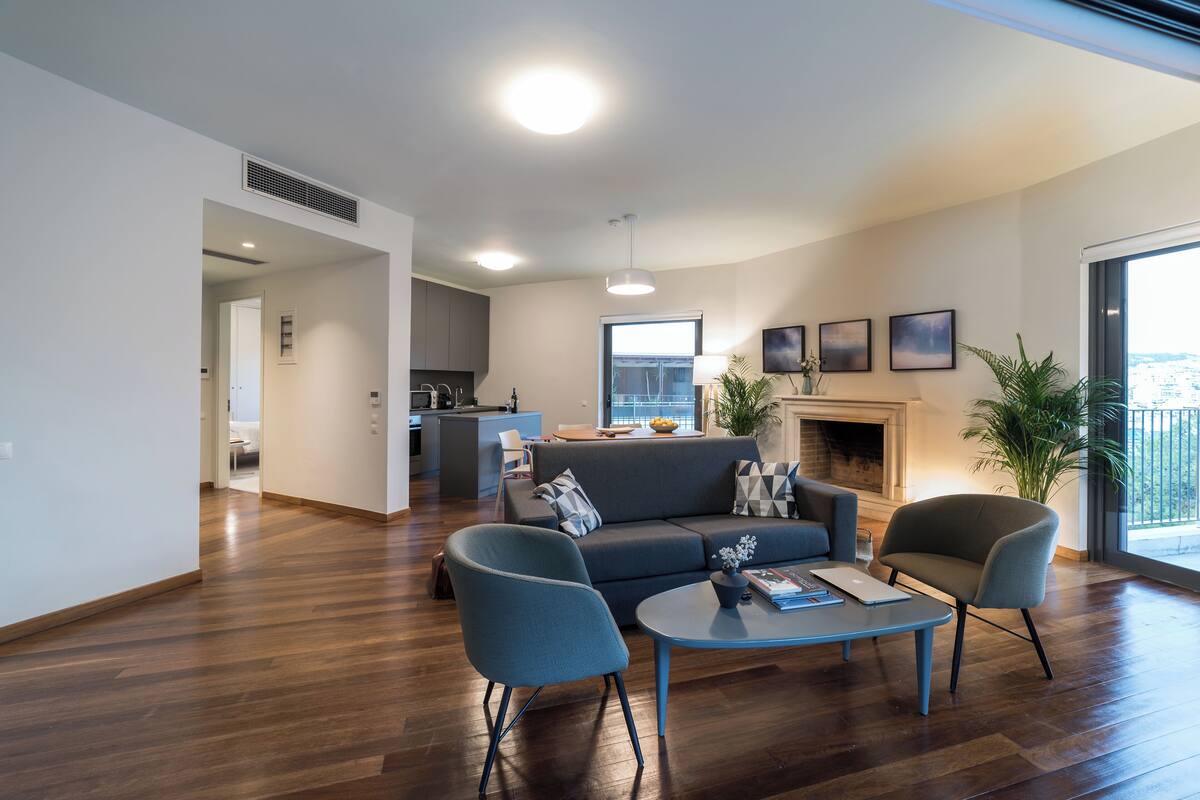 A modern living room with a sofa, two armchairs, and a coffee table. There is a fireplace, a kitchen in the background, and large windows.