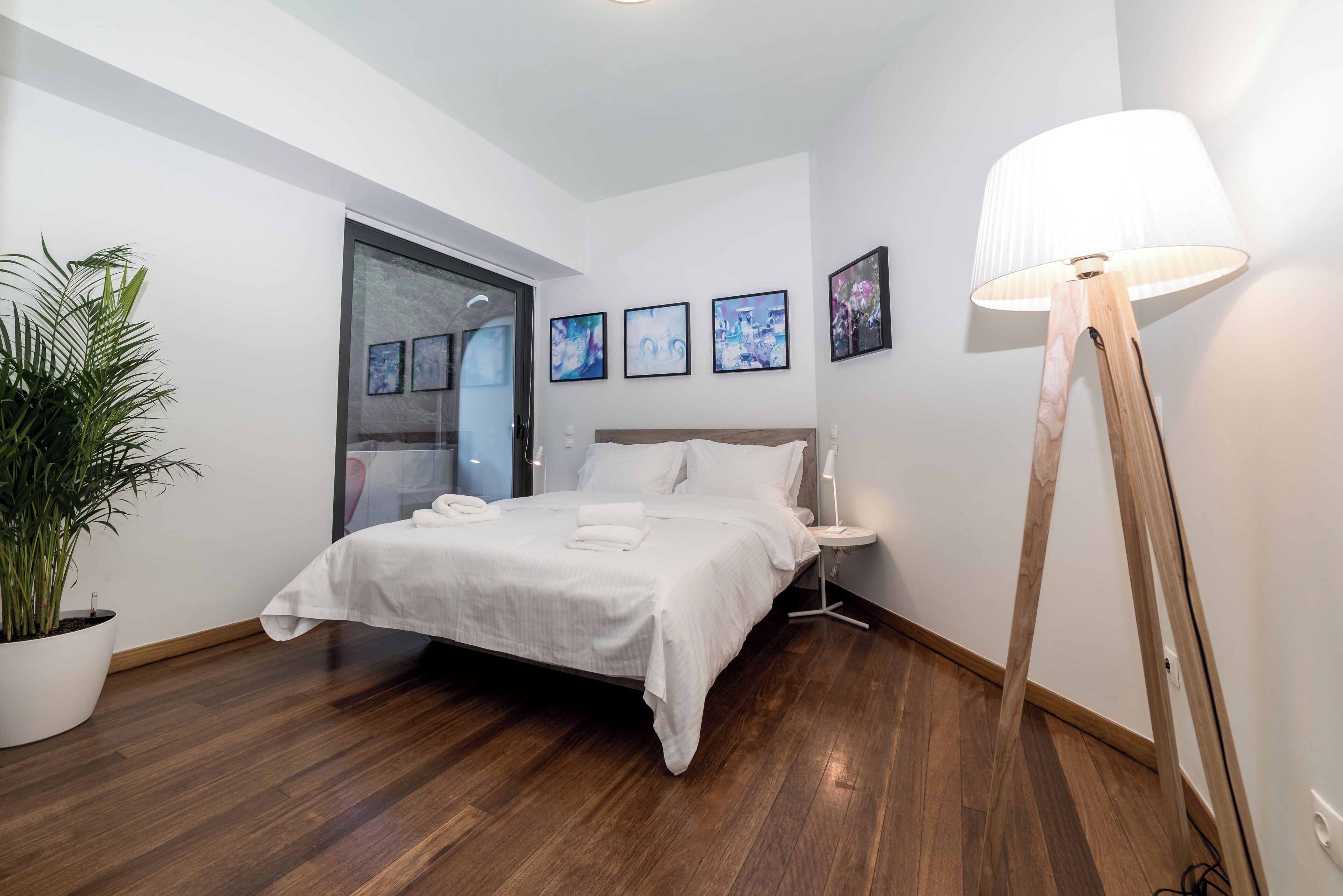 A modern bedroom with a large bed, a bedside table, a floor lamp, and a potted plant.