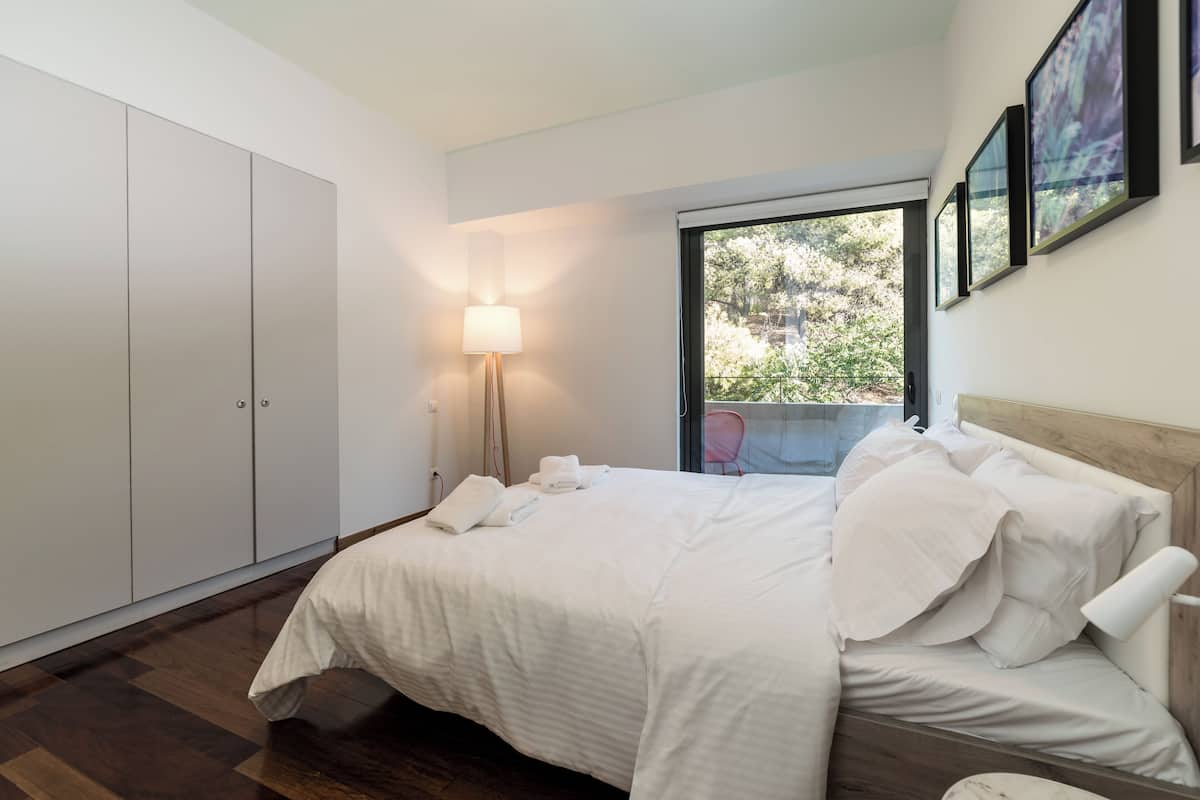 A bedroom with a large bed, a wardrobe, a bedside table, a lamp, and a window with a view of greenery.