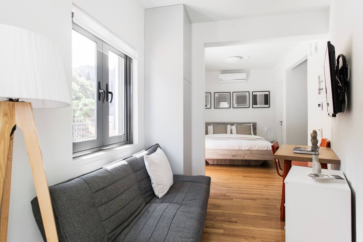 A modern, well-lit room with a grey sofa, a wooden table, a bed, and a small white fridge.