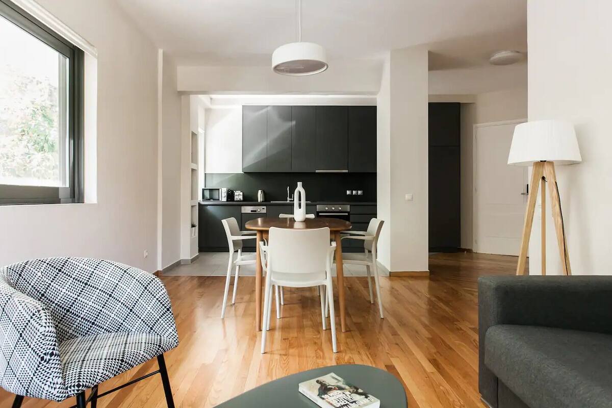 A modern living room with a dining area, a kitchen with dark cabinets, and a large window.