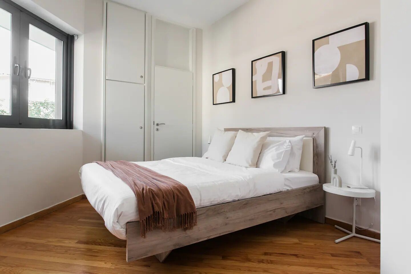 A bedroom with a wooden bed, white bedding, a bedside table with a lamp, and a window with blinds.