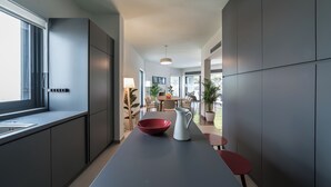 A modern kitchen with a long countertop, a red bowl, and a white pitcher. There is a dining area with a table and chairs in the background. - The Hill by Zoia Living (Athens)