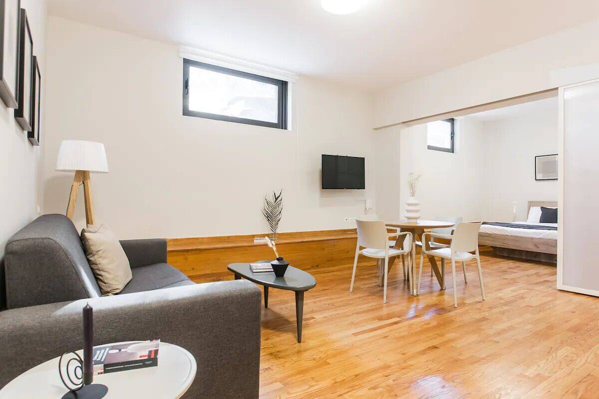 A modern hotel room with a sofa, a small table, a dining area with chairs, a bed, and a TV mounted on the wall.