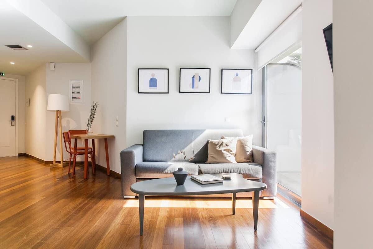 A modern living room with a grey sofa, a coffee table, and a dining area with a table and chairs. There are framed pictures on the wall and a large window providing natural light.