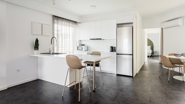 A modern kitchen with white cabinetry, a central island with bar stools, and a dining area with a table and chairs. - OneZeroTwo by Zoia Living (Athens)