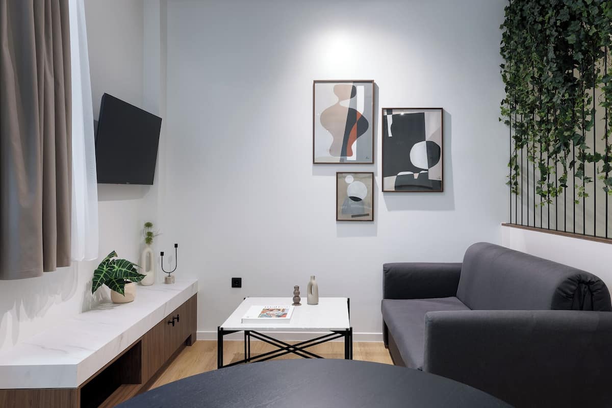 A modern living room with a grey sofa, a white coffee table, and a TV mounted on the wall. There are abstract paintings on the wall and a plant on the right side.