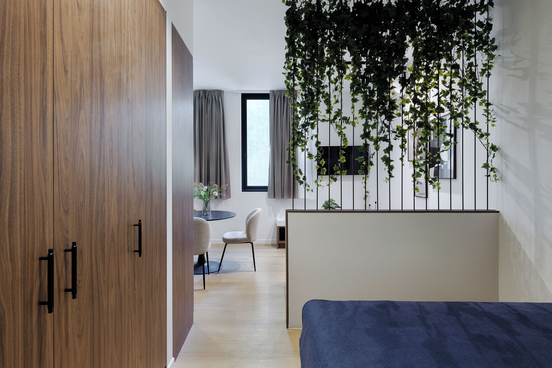 Une chambre moderne avec une armoire en bois, un lit recouvert d’une couverture bleue, une chaise, une petite table ronde et une fenêtre avec des rideaux.