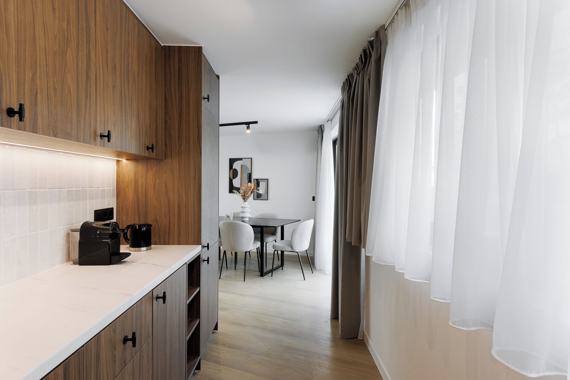 Une cuisine moderne avec des meubles en bois, un plan de travail sur lequel se trouve une machine à café, et un coin repas avec une table et des chaises.