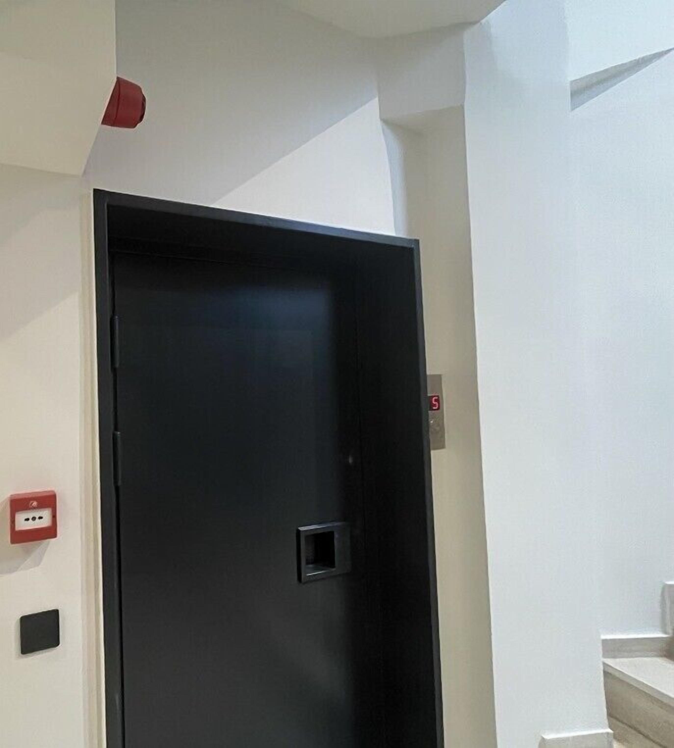 A black door with a handle, a red emergency exit sign, and a power outlet on the wall.