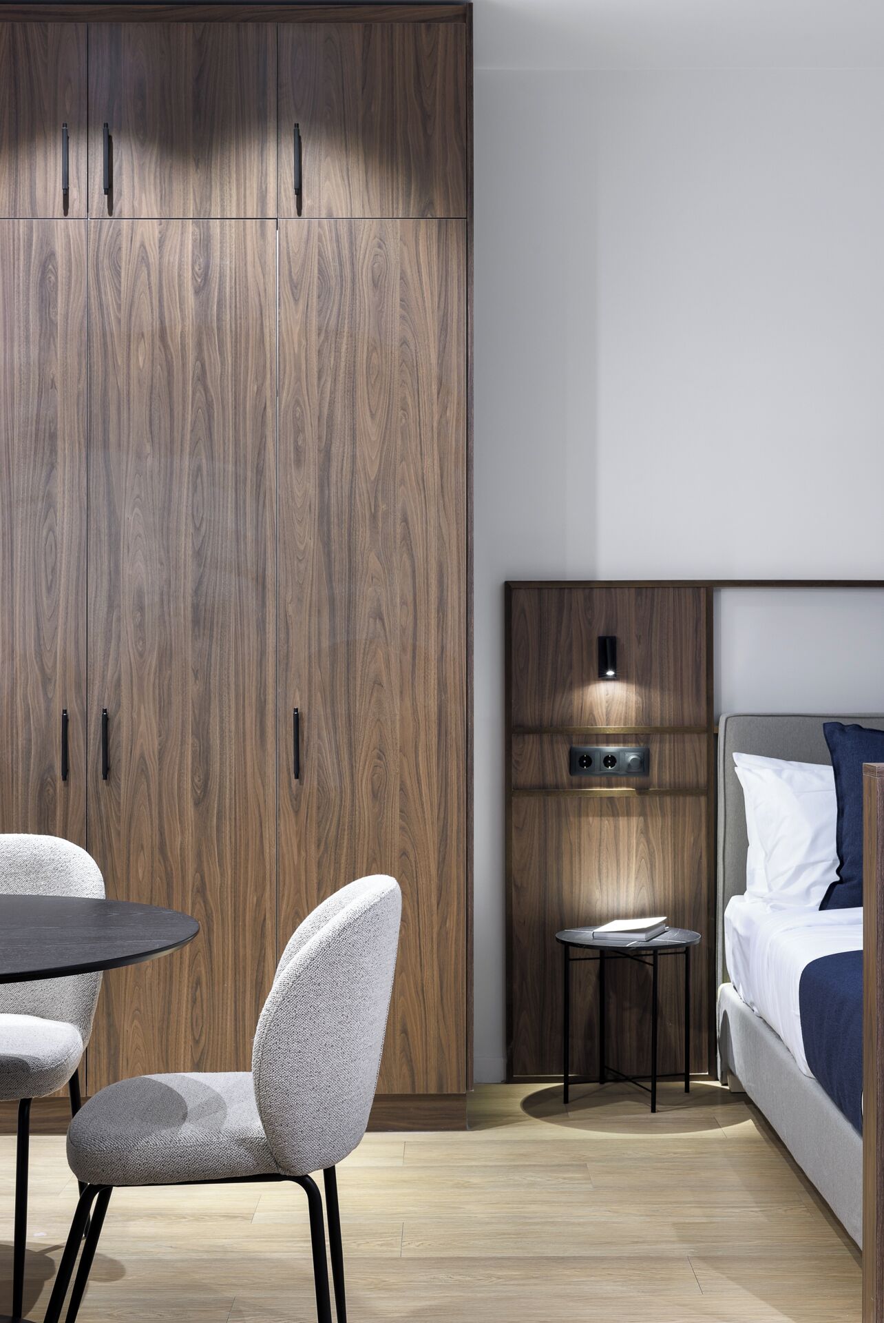 Una habitación de hotel moderna con un armario de madera, una mesa redonda con sillas, una mesita de noche con lámpara y una cama con almohadas.