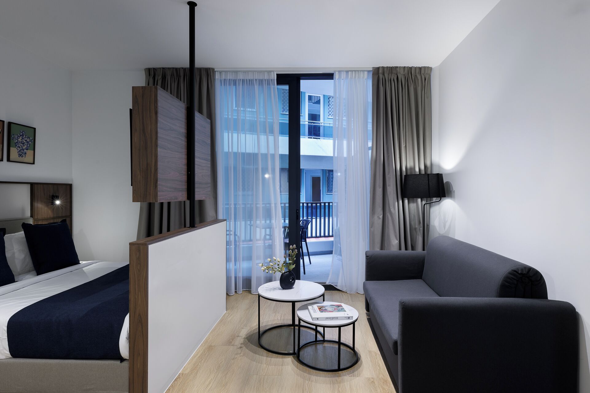 Et moderne hotelværelse med seng, sofa, et lille rundt bord og udsigt til en bygning gennem lette gardiner.