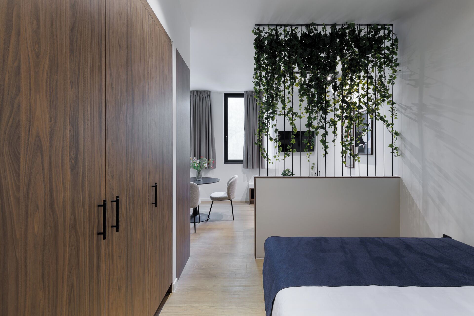 A bedroom with a wooden wardrobe, a bed with a blue cover, a desk with a chair, and a wall-mounted TV.