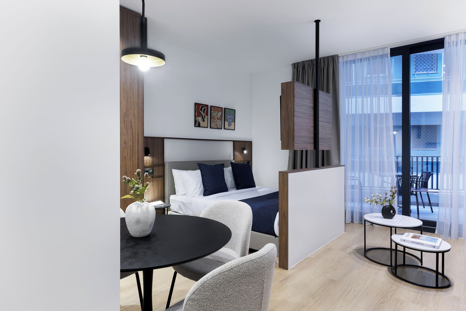 A modern hotel room with a bed, a dining table, a small side table, and a balcony with a view.