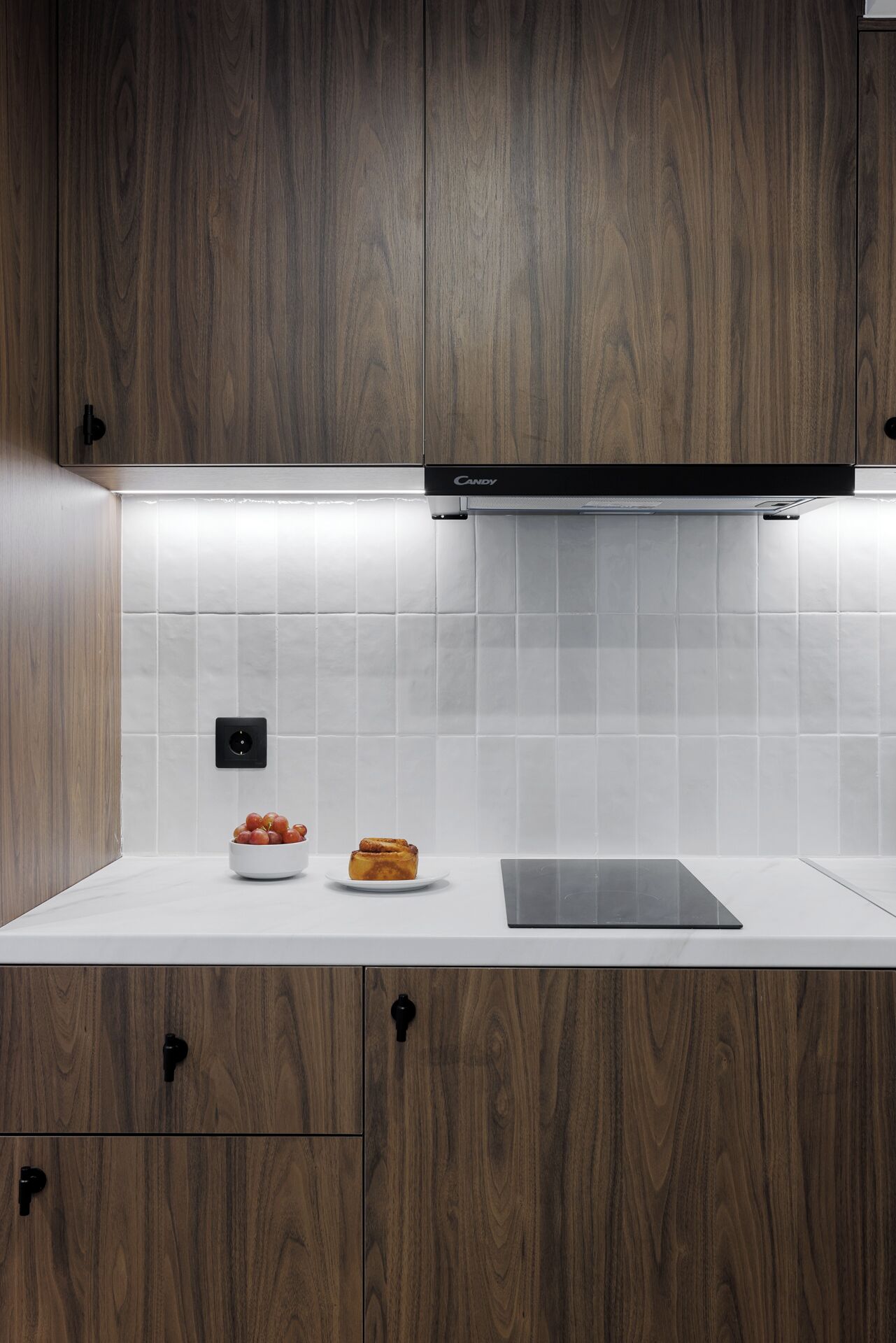 Une cuisine moderne avec des meubles en bois, un plan de travail blanc, une hotte noire et un pare-feu en carreaux.