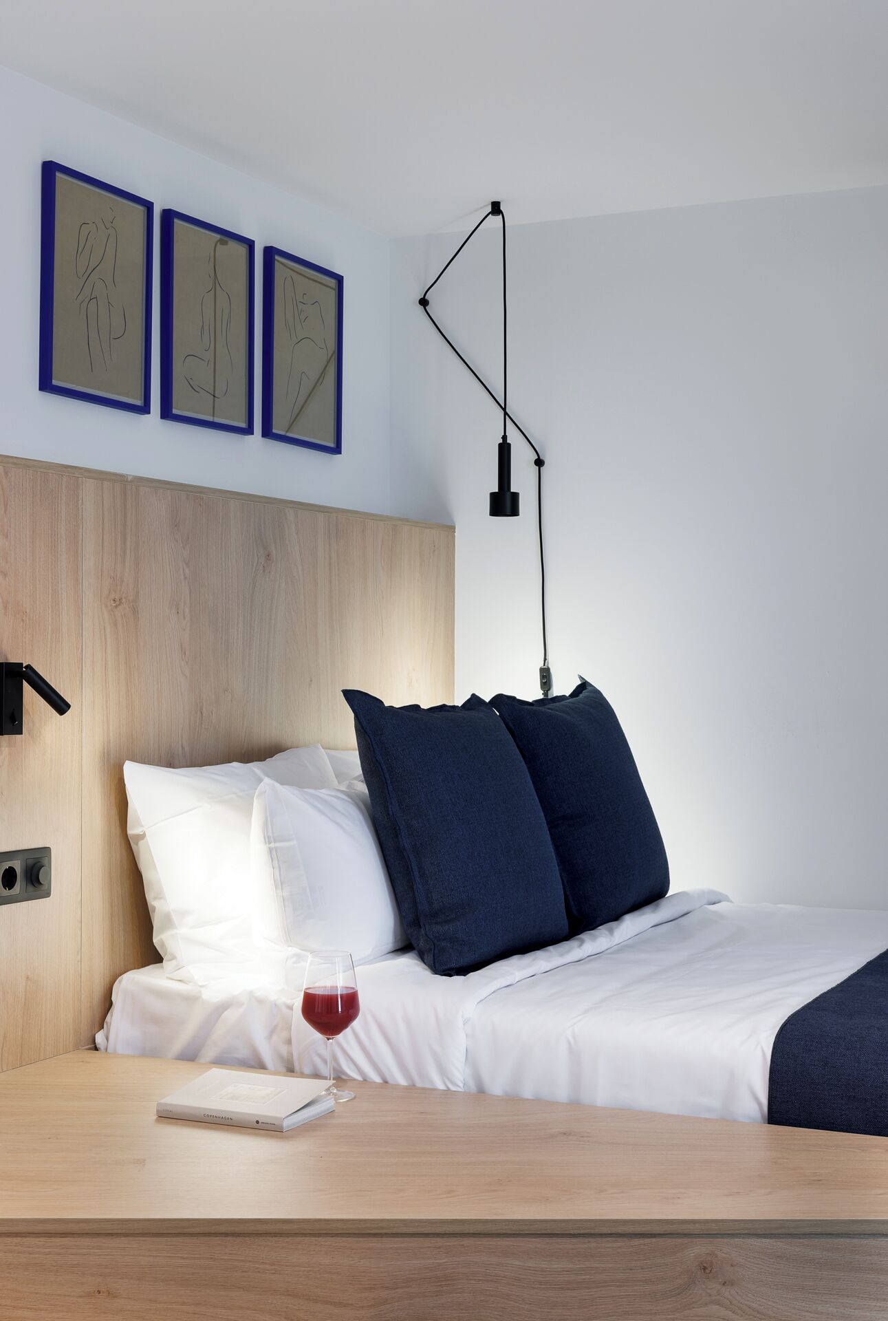 A neatly made bed with white and navy blue pillows, a bedside table with a glass of red wine, and three framed artworks on the wall.