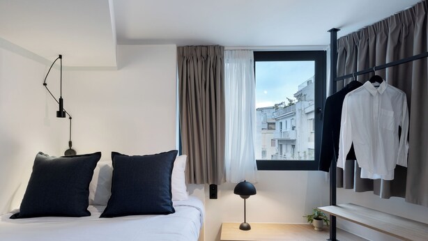 Ein modernes Hotelzimmer mit Bett, Nachttisch, Lampe und Blick auf die Stadt durch ein Fenster.