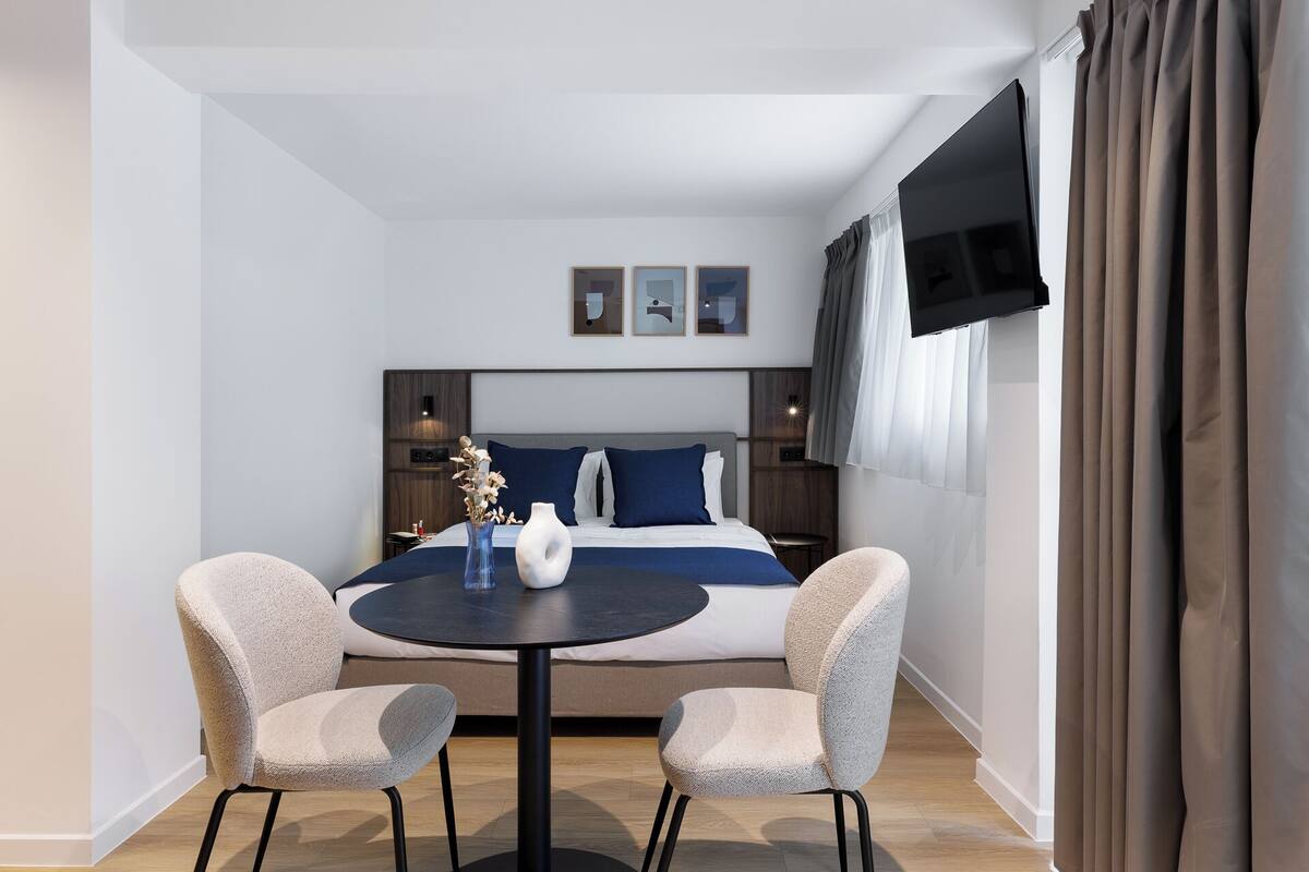 A modern hotel room with a bed, two armchairs, a small round table, and a TV.