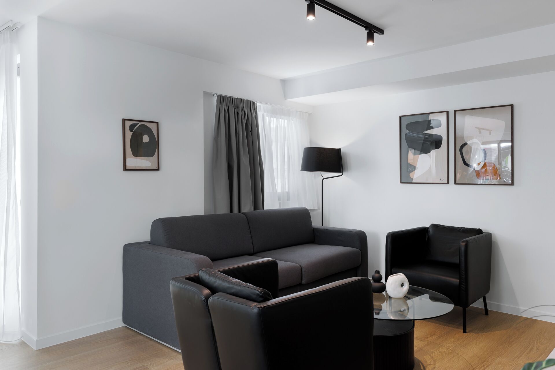 A modern living room with a grey sofa, two black armchairs, a glass coffee table, and a floor lamp.
