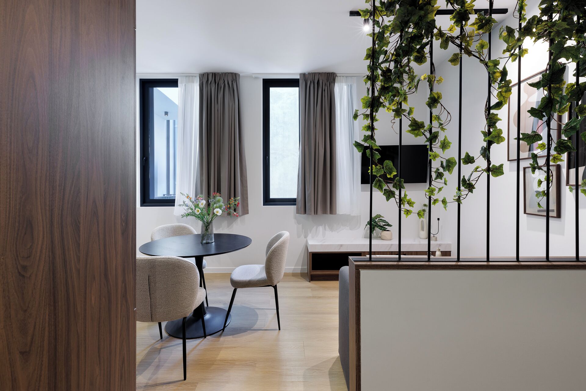A modern living room with a round table, two chairs, a TV mounted on the wall, and a large window with curtains.