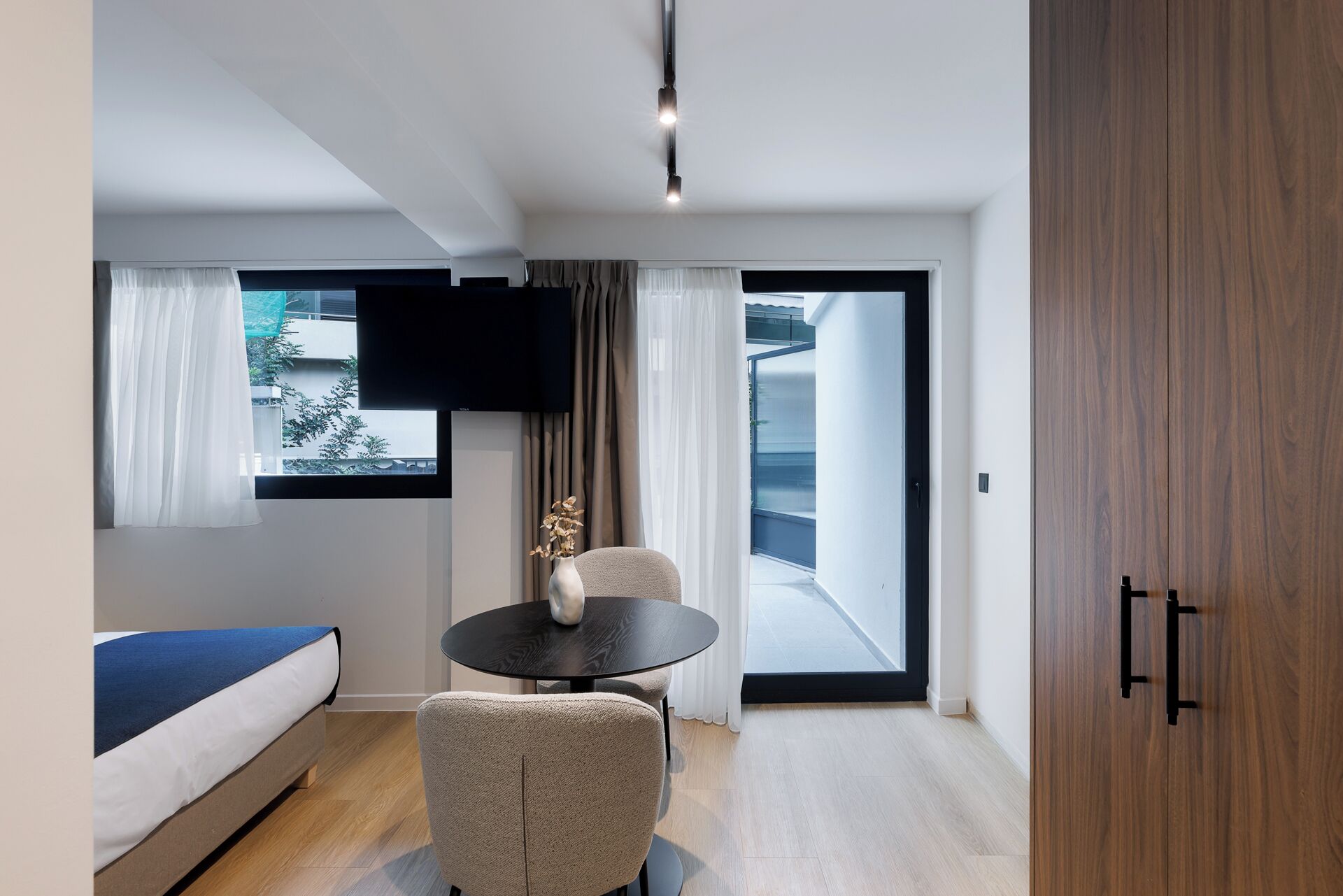 Ein modernes Hotelzimmer mit einem Bett, einem runden Tisch mit einer Vase, einem Stuhl, einem an der Wand befestigten Fernseher und einer Glastür, die auf einen Balkon führt.