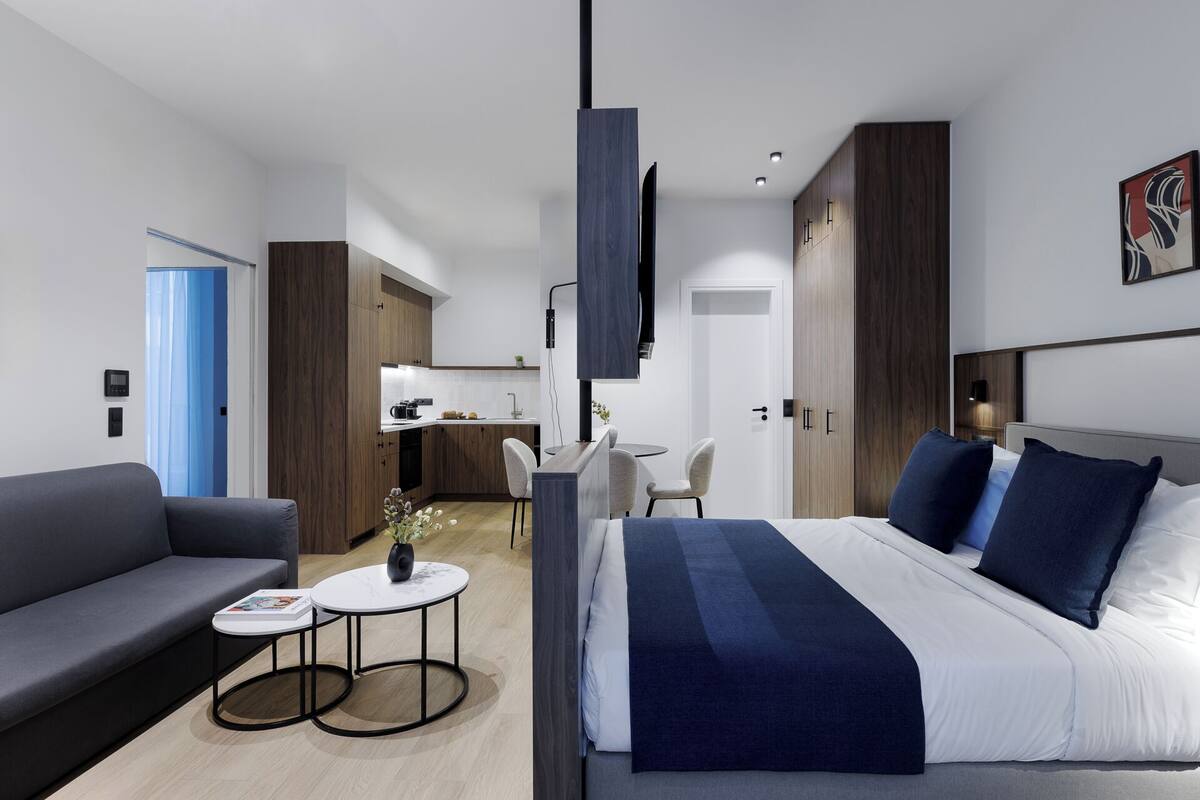 A modern hotel room with a large bed, a sofa, a dining area, and a kitchenette.