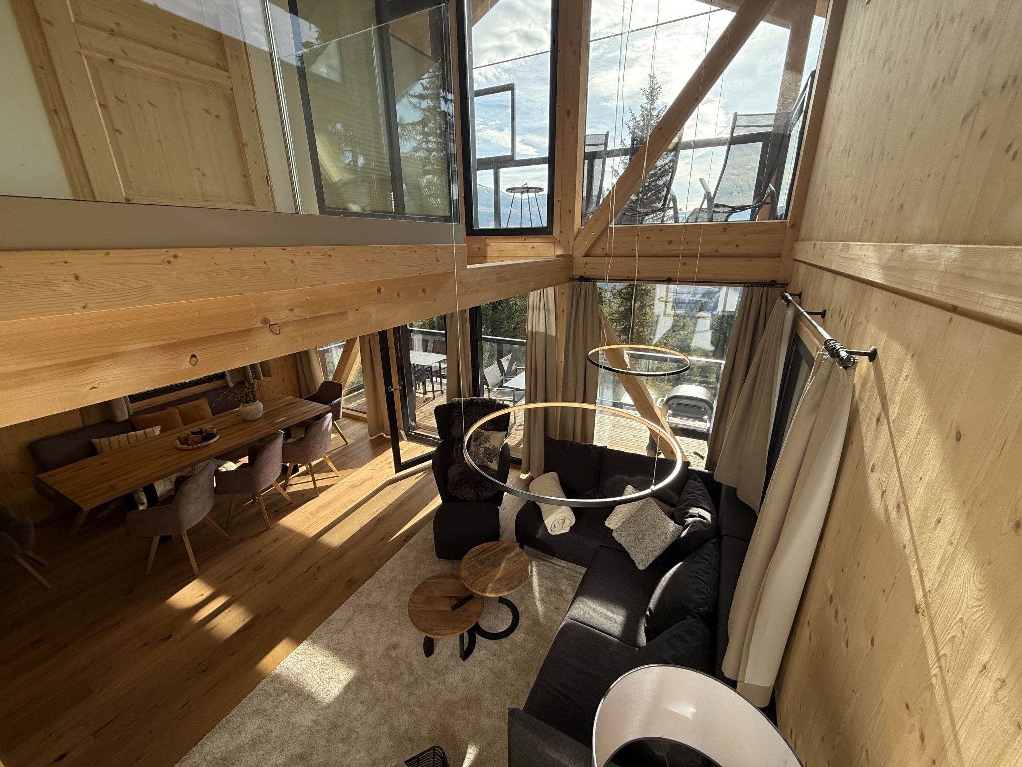 Um interior moderno com teto alto, vigas de madeira, área de jantar com uma mesa comprida e cadeiras, um sofá e um lustre circular.