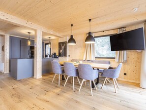 Een modern interieur met een eethoek van hout, gestoffeerde grijze stoelen en een flatscreen televisie aan de muur.