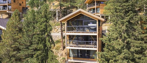 Una cabaña de madera de varios pisos con ventanas grandes, rodeada por un bosque frondoso.