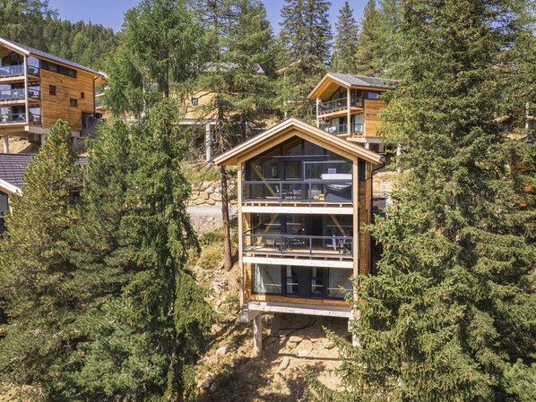 A multi-story wooden cabin with large windows, surrounded by dense forest.